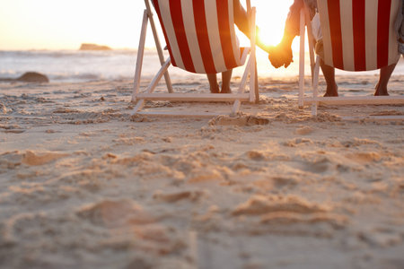 Couple, back and holding hands on beach for travel, bonding and holiday on anniversary date with sunshine. Man, woman and relax by ocean for trip, love and tropical vacation with chair in Maldivesの写真素材
