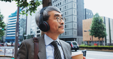 Mature businessman, headphones and thinking for street, commute and music with subscription on Tokyo trip. Japanese person, drink and walking to workplace with podcast, streaming and urban japan roadの写真素材