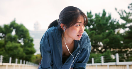 Tired, woman and fitness with earphones in city from sport exercise, marathon training and listen to music. Athlete, fatigue and break of cardio workout, physical recovery and podcast on Japan bridgeの写真素材