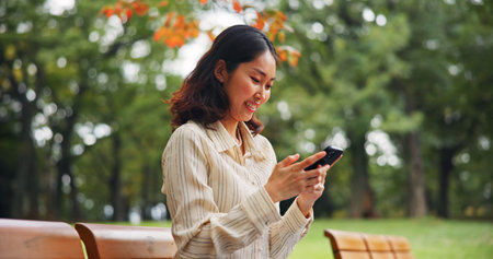 Woman, relax and typing with phone at park for travel location, communication and directions guide. Smile, person and mobile on vacation, social media and information with commute navigation in Japanの写真素材