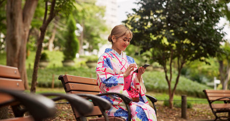Smile, park and Japanese woman with phone for social media, text message or reading ebook on internet. Asian girl, mobile and outdoor on bench for relax, chat app and traditional kimono for cultureの写真素材