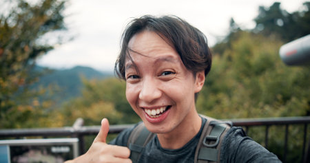 Man, thumbs up and outdoor in portrait for selfie, fitness update and follower interaction on Mount Takao. Japanese influencer, like emoji or smile on bridge for travel, hiking or social media streamの写真素材