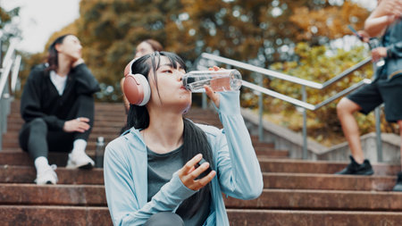 Woman, headphones and drinking water for training in park, mineral liquid and fitness recovery. Female person, athlete and nutrition for hydration on stairs, workout break and streaming song in Japanの写真素材