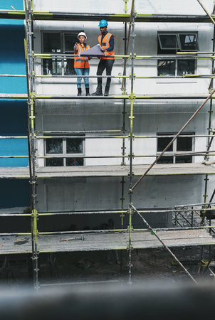 Blueprint, man and woman on construction scaffolding with planning, collaboration and urban architecture. Ideas, discussion and contractor team on site for civil engineering, building and developmentの写真素材