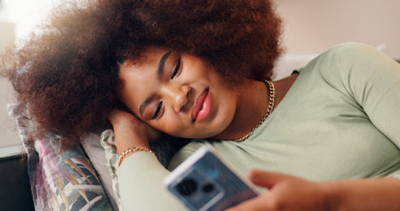 Smile, mobile and woman in home online, texting and reading social media blog post to relax. Happy, phone and African girl on sofa for communication, streaming show and download chat app on internetの写真素材