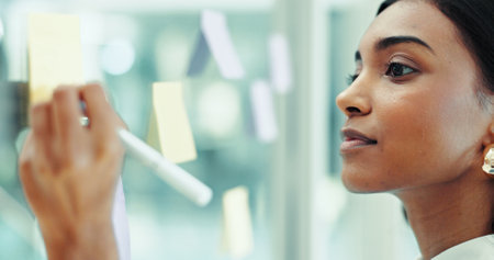 Glass wall, writing and woman in office for planning, brainstorming or research for creative project. Journalism, thinking and female editor working with notes for media report for startup business.の写真素材