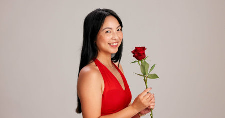 Woman, studio background and happy with rose on portrait for love, care and valentines day celebration in Japan. Japanese person, romance and smile in red dress with flower as anniversary giftの写真素材