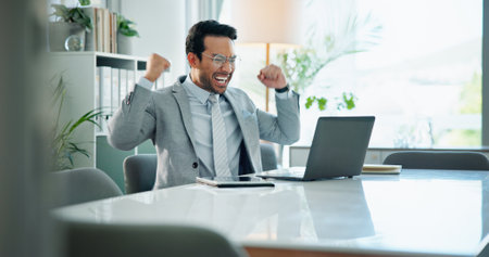 Office, happy man and success with celebration, laptop and business and investment banking firm. Male person, victory and technology in communication for financial review, audit or career opportunityの写真素材
