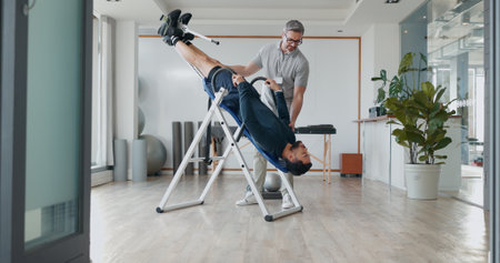 People, inversion table and upside down for physical therapy with support, balance and recovery at clinic. Physiotherapist, men and patient with fitness bench, biokinetics and rehabilitation processの写真素材