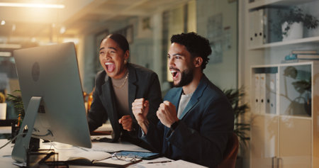 Business people, excited or computer in office for news, stock market win or promotion at night. Finance team, cheers or tech in firm for celebration, positive feedback or project success at deadlineの写真素材