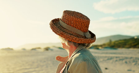 Senior woman, hat and view on beach for travel adventure, sightseeing and exploring nature in retirement. Back, elderly person and relax by ocean for fresh air, wellness and holiday break on weekendの写真素材