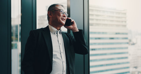 Japanese, businessman and office with phone call for communication, planning or networking by window. Thinking, male person and mature lawyer with mobile for talking, smile and case detail for clientの写真素材