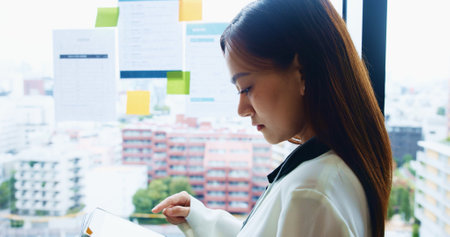 Office, ideas and woman at window with tablet, checking business plan or online schedule for workshop. Reading, scroll and Japanese businesswoman on digital app for report, research or notes on glassの写真素材