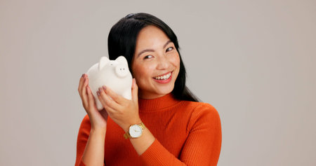 Woman, happy and listen to piggy bank in studio with laughing, saving and coins by background. Japanese person, girl and shake container for cash, money and excited with financial growth for wealthの写真素材