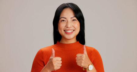 Happy, portrait and asian woman with thumbs up for thank you, good job or feedback on a studio background. Face, female person or model with smile, like emoji or yes sign for agreement or well doneの写真素材