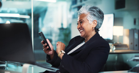 Mature, business woman and smartphone at desk for stock market research or checking investment account. Professional, night trader and smile with phone in office for economy news and trading scheduleの写真素材