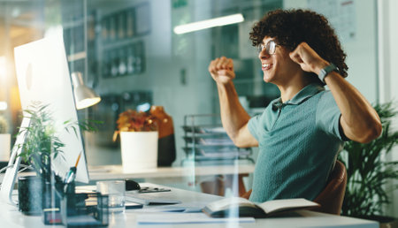 Happy, businessman or night with computer for winning, promotion or celebration at office desk. Excited, man or employee with smile or fist pump for victory, achievement or good news at workplaceの写真素材