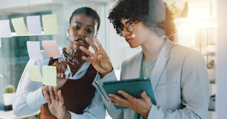 Man, woman and tablet by glass wall, notes and planning with teamwork, review and project at startup. People, sticky note and touchscreen with app, discussion and brainstorming at creative agencyの写真素材