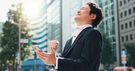 Businessman, phone and celebration in city for success, client acquisition and development deal. Japanese person, mobile and fist bump outdoor for financial profit, investment growth and achievementの写真素材