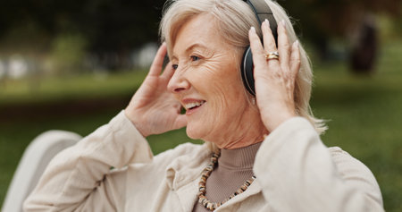 Senior woman, headphones and relax with music at park for audio streaming, podcast or radio in nature. Mature, female person or retirement with earphones for sound, listening or outdoor entertainmentの写真素材