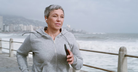 Running, health and music with old woman at beach for fitness podcast, wellness and streaming. Athlete, training and workout with senior person and earphones for challenge, exercise and cardioの写真素材