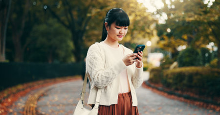 Japanese woman, walking and phone in park for online networking, dating app or travel with mobile chat. Person, smartphone and smile for social media post, notification or browsing internet in streetの写真素材