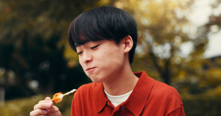 Street food, park and Japanese man with eating, smile and asian snack on trip outdoor. Happy, dumpling and hungry student on summer break and vacation in Tokyo with lunch and traditional cuisineの写真素材