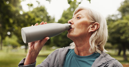 Mature, woman and drinking water in park for fitness, health and wellness in nature. Hydration, thirsty and bottle for electrolytes with exercise, self care and senior female person outdoor for breakの写真素材