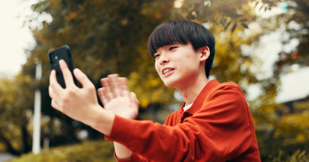 Phone, video call and man in park with wave, outdoor live streaming and online communication. Smartphone, influencer or Japanese streamer in nature for content creation, networking or connectivityの写真素材