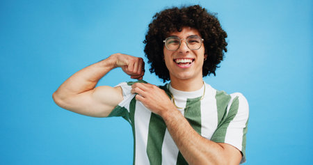 Studio, man and portrait with bicep for flex, muscle growth and development with smile for progress. Happy, male person or bodybuilder with pride for arm strength, wellness results or blue backgroundの写真素材