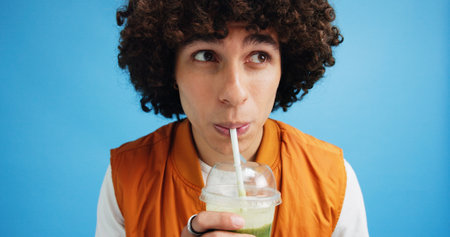 Studio, man and wellness with smoothie for drinking, digestive health and taste juice with thinking. Thoughts, male nutritionist and detox with green shake for diet, nutrition and blue backgroundの写真素材