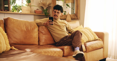 Man, phone and texting on sofa, relax and reading in living room, chat and mobile app in home. Person, smartphone and check notification with contact on lounge couch with social network at apartmentの写真素材