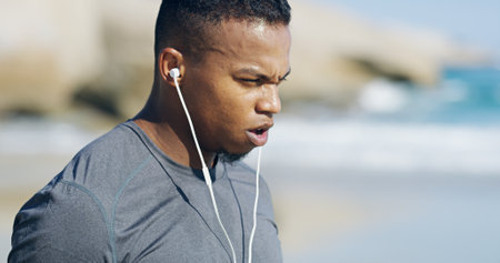 Beach, man and tired with breathing for fitness, exercise and sprinter with workout music for wellness outdoor. Ocean, male person and runner with sweat for training, cardio and earphones for podcastの写真素材