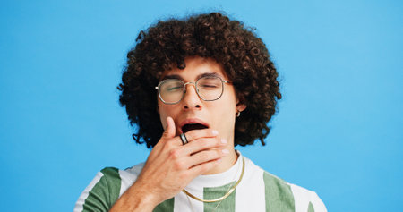 Yawn, tired and man in studio with fatigue expression, emoji or gesture. Geek, bored and afro guy with glasses and burnout with exhausted, style or hand over mouth insult on blue background mockupの写真素材