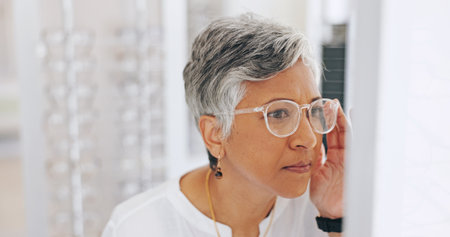Mature woman, glasses and choice in mirror at clinic for healthy vision, wellness and optometry services. Person, client and customer with decision for prescription eyewear, sight and frame at storeの写真素材