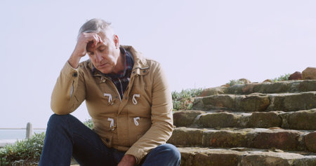 Senior, man and headache with stress on stairs for heat stroke, burnout or fatigue by ocean coast. Mature, male person or tired with migrine, pain or ache for mistake or vertigo on steps in natureの写真素材