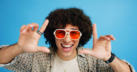 Happy man, portrait or sunglasses with hairstyle or funky fashion on a blue studio background. Male person, hipster or young model with stylish shades or finger frame for summer party or festive moodの写真素材