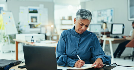 Office, laptop and mature woman with notes, business plan and online website project for graphic design. Schedule, research and businesswoman writing creative ideas in notebook at digital agency.の写真素材