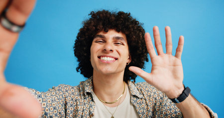 Selfie, wave or portrait of happy man in studio with hand gesture, video call or fashion clothes. Smile, greeting or influencer on blue background for social media streaming, photo or vlogging povの写真素材