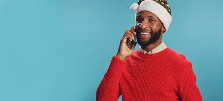 Christmas, man and phone call in studio with smile, good news and wishes for festive season in banner. Person, happy and Santa hat on blue background with contact, connection and chat at mockup spaceの写真素材