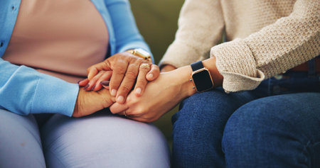 Senior woman, friend and holding hands for empathy in home with grief, support and compassion for loss. Sympathy, people and closeup of comfort on sofa for care, wellbeing and help with mental healthの写真素材