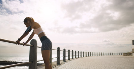 Seaside, woman and retired from running with fitness on earphones for wellness, health and self care. Female person, cloudy sky and space on break with jogging as workout, training and exerciseの写真素材