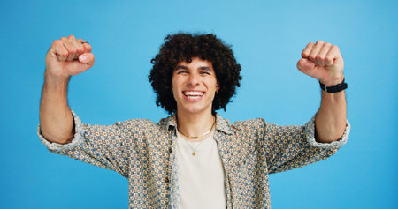 Happy man, portrait and winning with fist pump for celebration or promotion on a blue studio background. Excited, male person or young model with smile in joy for victory, prize or holiday dealの写真素材