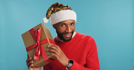 Portrait, black man and gift for Christmas in studio of festive celebration, surprise present and Santa Claus. Smile, male person and box for giveaway announcement, holiday reward and blue backgroundの写真素材