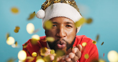 Christmas, man and blow confetti in portrait for celebration, festive spirit and excited in studio. Person, Santa hat or glitter on blue background for holiday event, Xmas party and culture in Brazilの写真素材