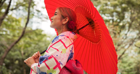 Park, umbrella and woman with fashion, thinking and travel with smile in woods. Forest, kimono and parasol with walking, outdoor and journey with morning, style and traditional Asian dress in natureの写真素材