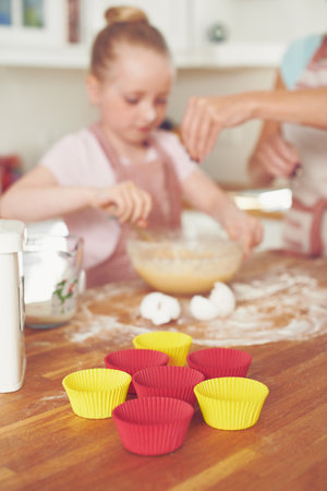 Mother, girl and cups in kitchen for baking, teaching and bonding with support, bowl and ingredients. Family, kid and help in house for cooking, cupcakes and development with flour, wheat or learningの写真素材