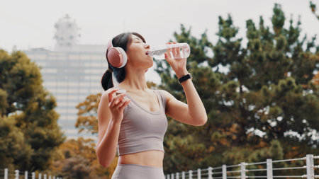 Drinking water, outdoor and woman with fitness, wellness and rest with workout. Japanese person, runner and athlete in city, headphones and listening to music, healthy and thirsty with hydrationの写真素材