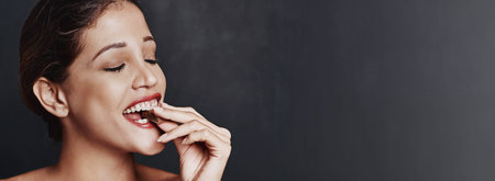 Happy woman, chocolate and bite with candy in studio for unhealthy diet, sweet or dessert on a dark...