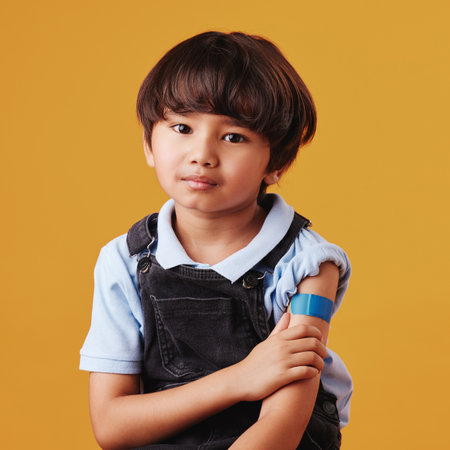 Asian child, portrait and plaster on studio background for vaccine, healthcare and illness prevention. Japanese boy, pain and bandage on arm for flu shot, immune system strength and first aid helpの写真素材